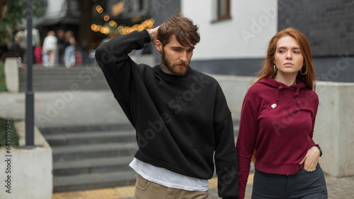 Fashion beautiful young woman model in fashionable urban outfit with hoodie walking together in the city. Pretty redhead young woman and handsome bearded hipster man in black sweatshirt © alones