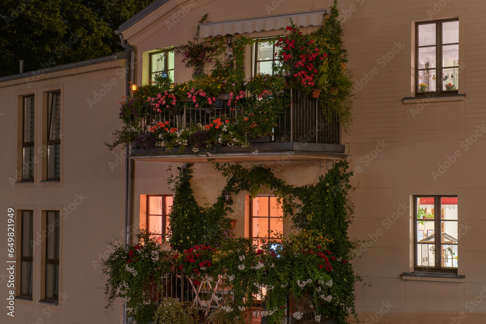 old house at night with light in the windows, balconies decorated with flowers and plants.