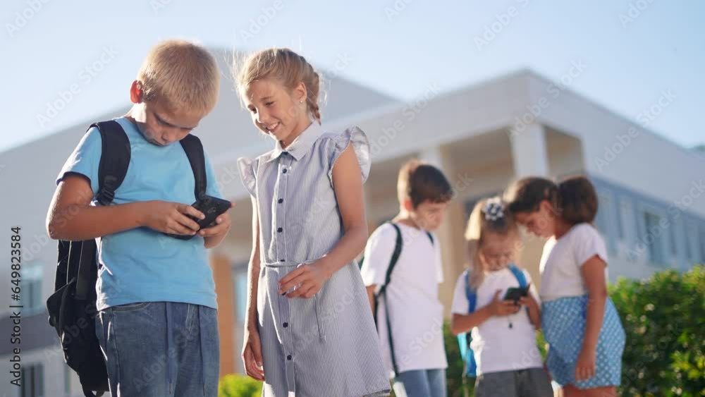 children group school children look at the smartphone video. school learning kid concept. group of kids with backpacks playing smartphone near the school building outdoors sunlight children group school children look at the smartphone video. school learning kid concept. group of kids with backpacks playing smartphone near the school building outdoors sunlight