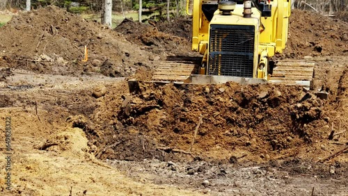A yellow Bulldozer pushes brown soil toward camera as it levels the ground in preparation for the building of a new home.