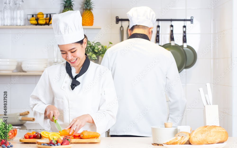 Two Asian professional couple chef wearing white uniform, hat, helping each other, preparing meal,cooking in kitchen together, smiling with happiness and confidence. Food Concept.
