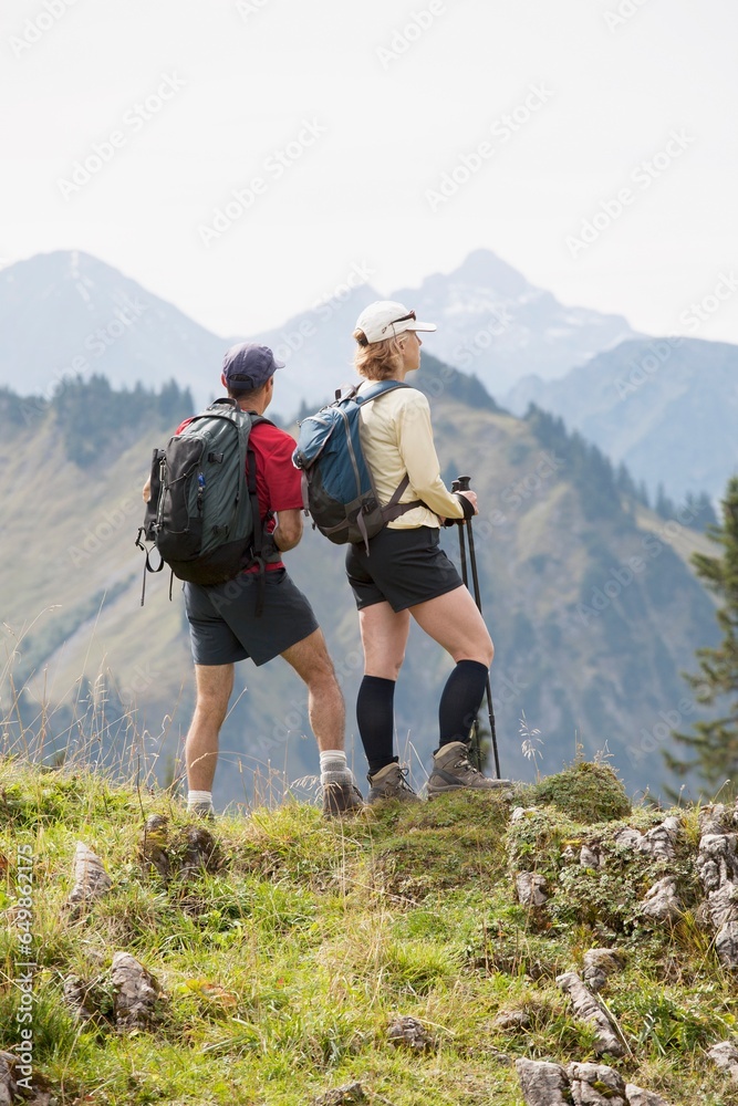 Female And Male Hikers On A Grassy Viewpoint Overlooking A Mountain Valley And Range; Oberstdorf, Germany