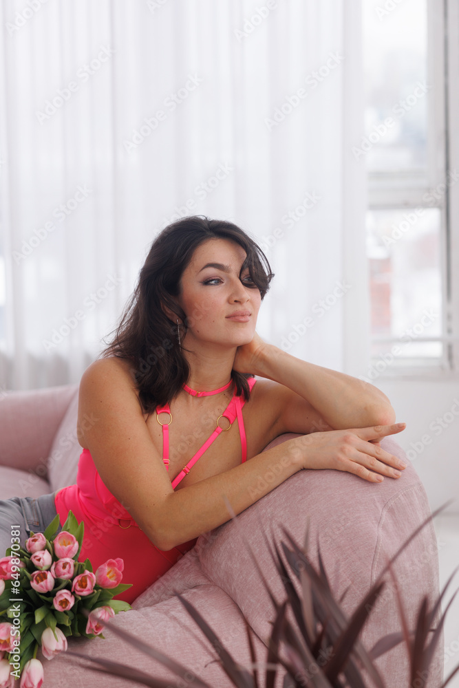 Beautiful brunette woman sitting on pink velvet sofa with a bucket of flowers tulips. Girl in pink bodysuit near window. Portrait of young woman smile. Living room.