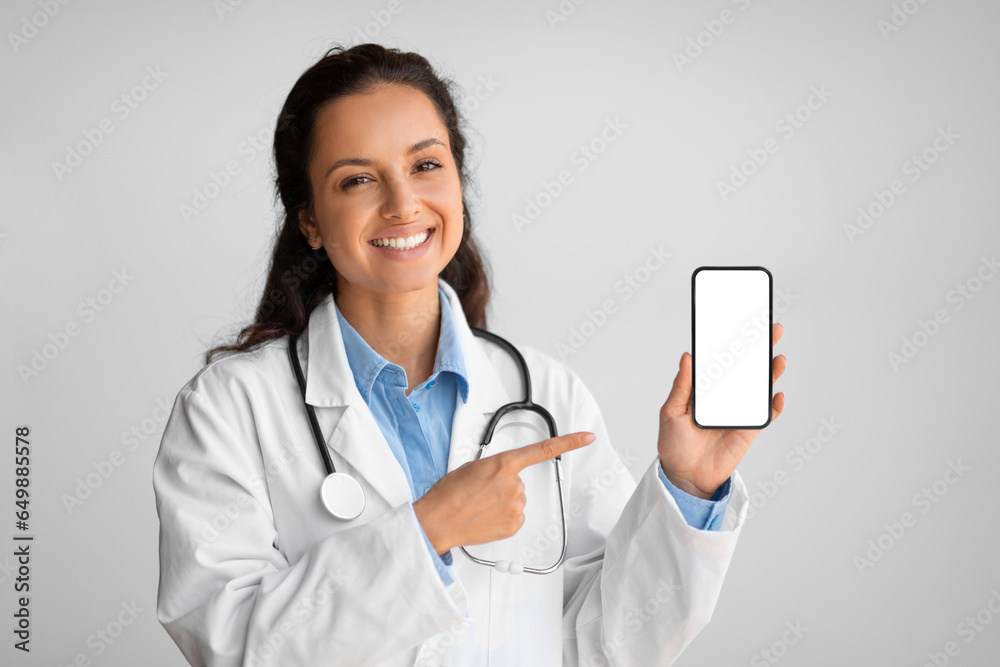 Smiling female physician showing and pointing at blank cellphone screen, standing on light wall background, mockup Smiling female physician showing and pointing at blank cellphone screen, standing on light wall background, mockup