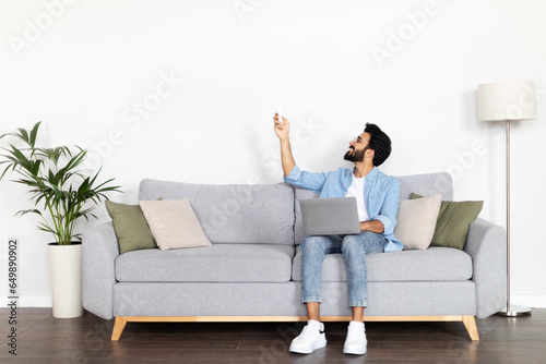 Happy indian guy freelancer sitting on couch, turning on AC