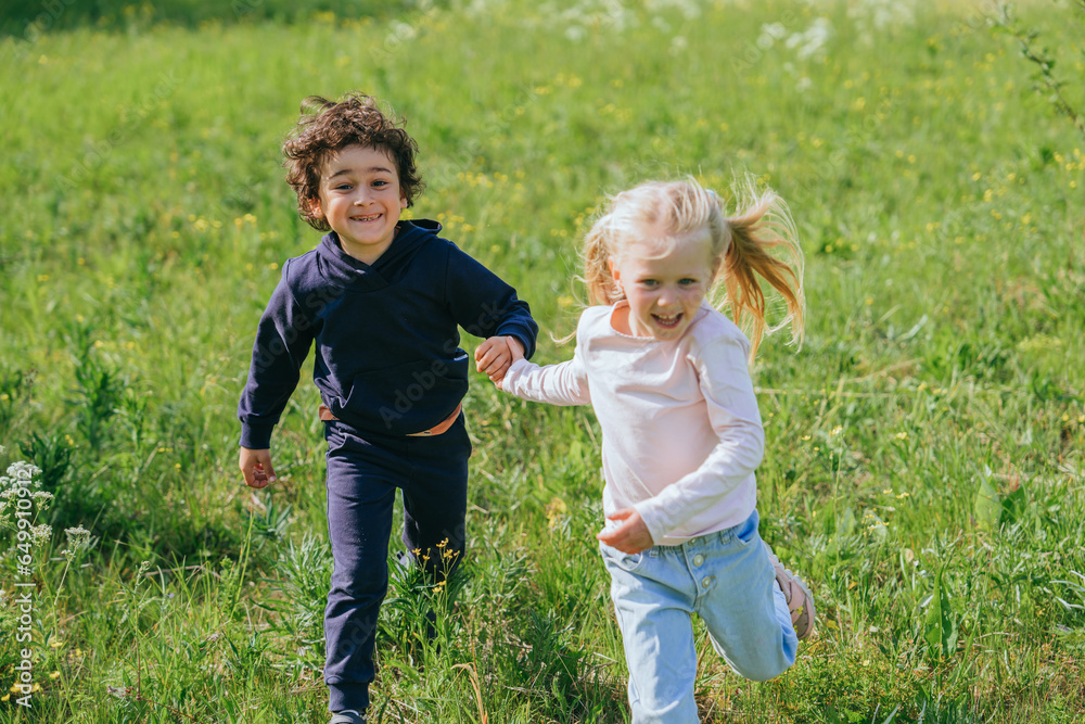 Cheerful brunette curly hispanic boy running at meadow with blonde caucasian little girl smiles wide on sunny summer day. Childhood outdoor leisure. Pretty Swedish girl holds brothers hand. Cheerful brunette curly hispanic boy running at meadow with blonde caucasian little girl smiles wide on sunny summer day. Childhood outdoor leisure. Pretty Swedish girl holds brothers hand.