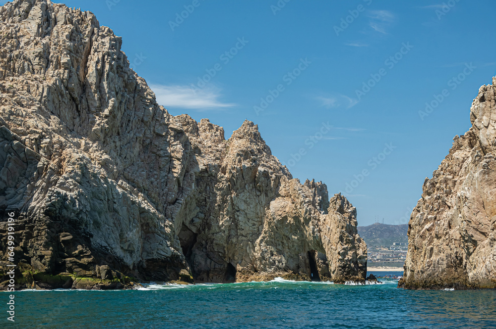 Mexico, Cabo San Lucas - July 16, 2023: South view on Reserva de Los Marina, open channel between tall gray boulders in greenish ocean water. Cityscape and beach on horizon