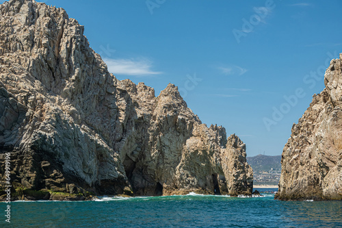 Mexico, Cabo San Lucas - July 16, 2023: South view on Reserva de Los Marina, open channel between tall gray boulders in greenish ocean water. Cityscape and beach on horizon © Klodien