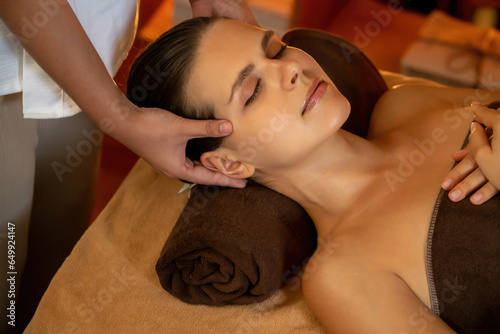 Caucasian woman enjoying relaxing anti-stress head massage and pampering facial beauty skin recreation leisure in warm candle lighting ambient salon spa in luxury resort or hotel. Quiescent