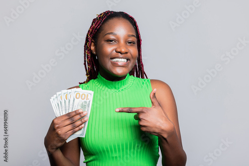 Delighted African American woman with colourful braids smiling holding cash dollars bunch of money in hands celebrating pointing with finger her first salary good payment photo