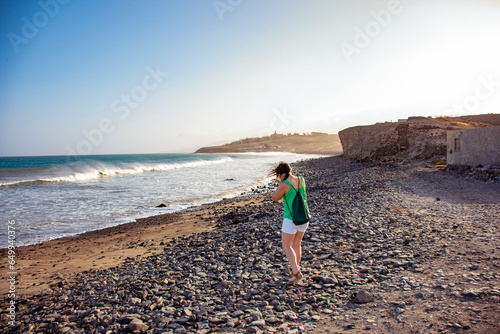 A white woman on her back with a drawstring backpack walking on the stones of the beach shore, careful not to trip over the wind blowing her hair.