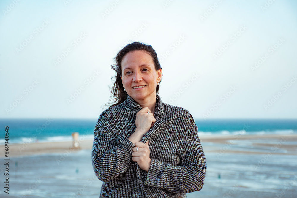 Mature white woman protecting herself from the wind with a linen jacket smiling on Sotavento beach in Fuerteventura with the beach in the background and the clear blue sky.