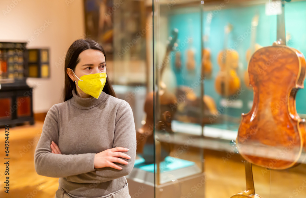 Female museum visitor in protective face mask examining with interest ancient stringed musical instruments displayed on exhibition. New normal in coronavirus pandemic - obrazy, fototapety, plakaty 