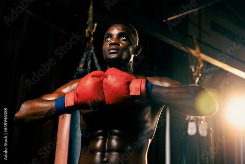 Boxing fighter posing, African American Black boxer put his hand or fist wearing glove together in front in aggressive stance and ready to fight at gym with kicking bag and boxing equipment. Impetus © Summit Art Creations