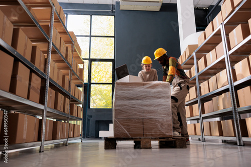 Diverse employees wearing industrial overall and helmet working in warehouse, preparing clients orders for delivery. Storage room workers checking shipping detalis on laptop computer. photo