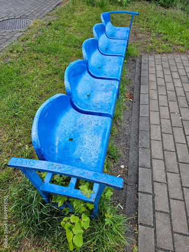 Картината върху платно A row of raindrop-covered blue plastic seats on the bleachers of a school football pitch on a drizzly summer day, Lodz, Poland