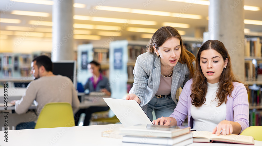 Obraz premium Two young women finding information at library together, working with laptop and books
