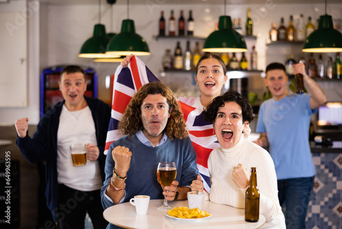 Company of joyful sports supporters waving flag of Great Britain and celebrating victory of favotite team in pub