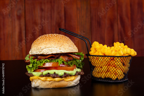 giant hamburger accompanied by an iron basket containing fries on a black table, with reflection and wooden background