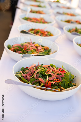 Photo of a long line of fresh green salads in white bowls on a white linen tablecloth for a gourmet dinner event.