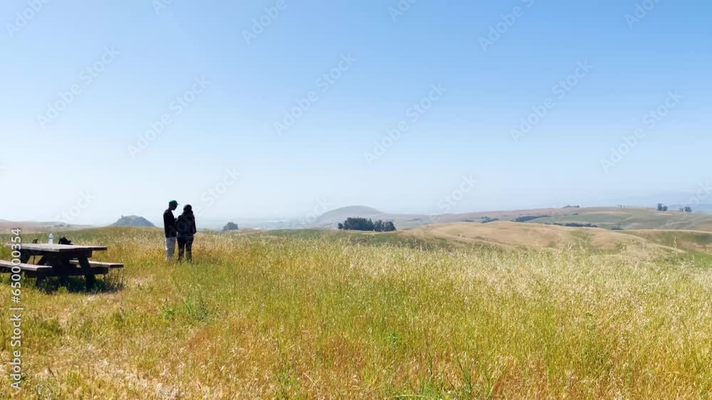 A pair of hikers walking through an open field under a clear sky. The panning shot captures the vastness and beauty of the natural landscape. A pair of hikers walking through an open field under a clear sky. The panning shot captures the vastness and beauty of the natural landscape.