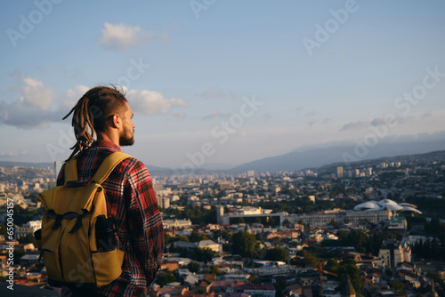 Beautiful view of the capital of Georgia Tbilisi from old town. A young man with a yellow backpack admires houses and skyscrapers. The guy is a tourist with dreadlocks. Rear view.