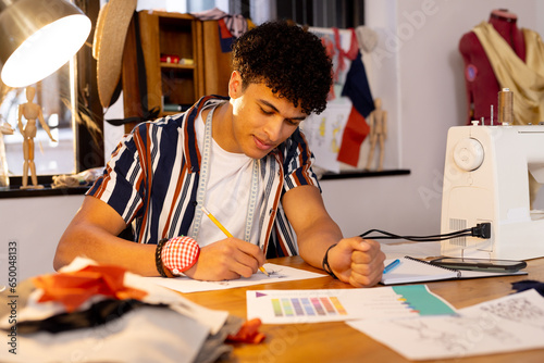 Focused biracial male fashion designer sketching designs and sitting at desk in studio photo