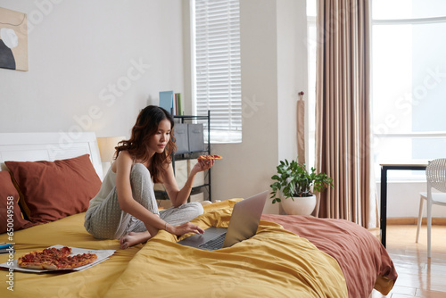Young woman sitting on bed, eating pizza and watching show on laptop