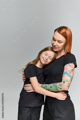 two generations, redhead woman and girl in matching attire hugging on grey backdrop, family love
