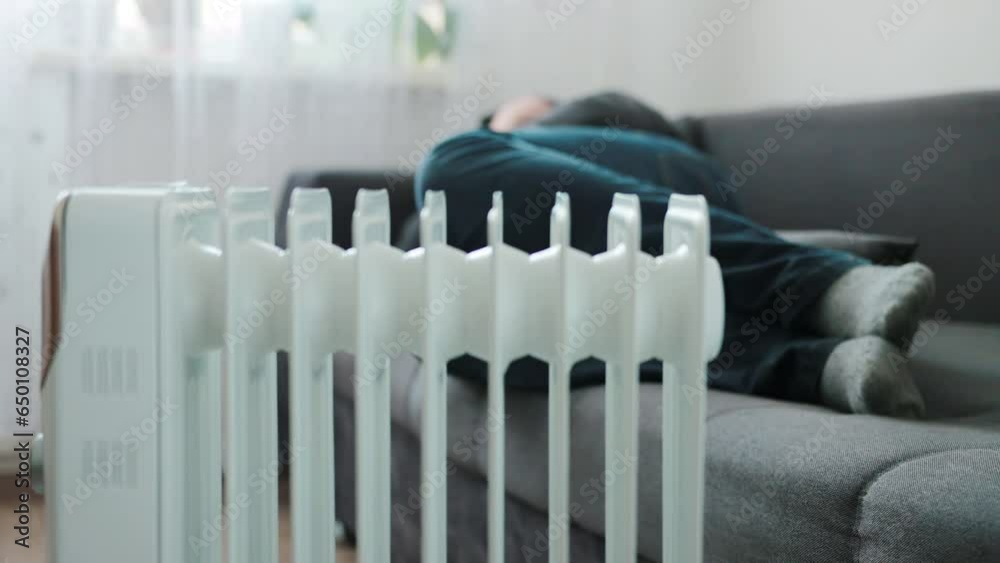 A man in a warm jacket with a hood lies on the sofa against the backdrop of a switched-on electric heater. The concept of heating a room in the off-season and cold times. Heat irradiation A man in a warm jacket with a hood lies on the sofa against the backdrop of a switched-on electric heater. The concept of heating a room in the off-season and cold times. Heat irradiation