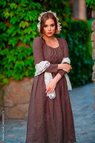 Beautiful red haired girl in brown long medieval dress and bonnet standing near stone wall. Art work of romantic lady.Pretty tenderness model posing on nature.