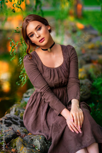 Beautiful red haired girl in brown long medieval dress and black vintage choker sitting on stone wall. Art work of romantic lady .Pretty tenderness model posing on nature.