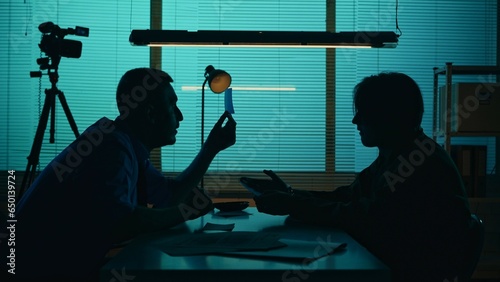 Medium silhouette shot of an offender, perpetrator or prisoner sitting in the interrogation room in front of the detective. The policemnan pushes on the suspect to get the confession.
