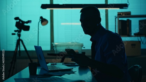 Medium silhouette shot of a detective, policeman sitting in the interrogation room, looking at his phone, working on a laptop and studying the evidence.