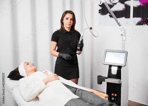 Fotografie Female cosmetologist holding radiofrequency device and looking at camera while standing by client in cosmetology clinic