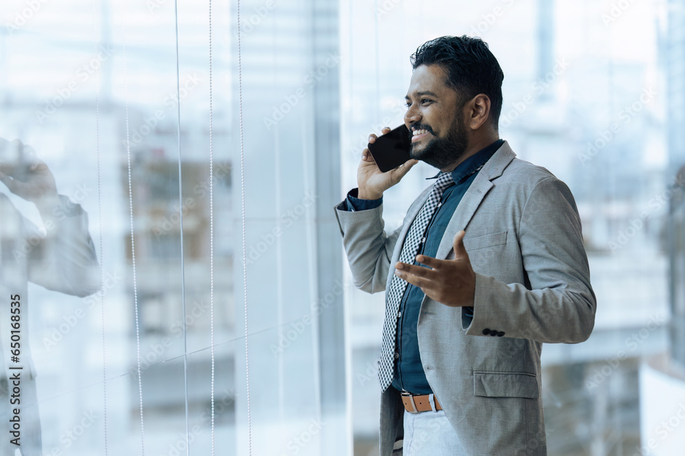 Indian businessman using smartphone talking having phone call conversation in office looking out window Indian businessman using smartphone talking having phone call conversation in office looking out window