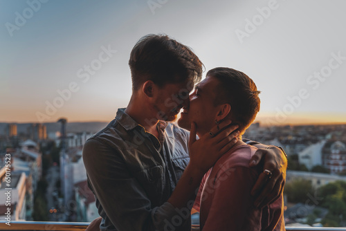 A young gay couple in love standing on a balcony overlooking the city at sunset and tenderly kissing © olezzo