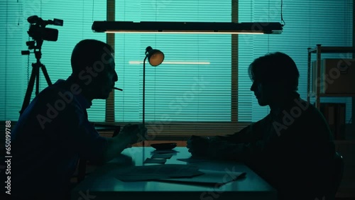 Medium silhouette video of an offender, perpetrator or prisoner sitting in the interrogation room in front of the detective. The policemnan argues with the suspect. photo
