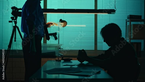Silhouette video of a perpetrator or prisoner sitting in the interrogation room in front of the detective. The policemnan pulls a gun from evidence box, talks to the suspect. photo