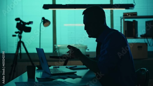 Medium silhouette video of a detective, policeman sitting in the interrogation room, smoking, working on a laptop and studying the evidence. photo