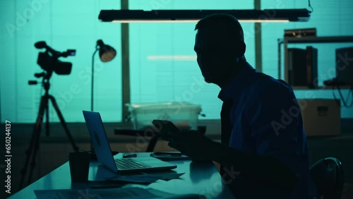 Medium silhouette video of a detective, policeman sitting in the interrogation room, smoking, working on a laptop and talking on the phone. photo