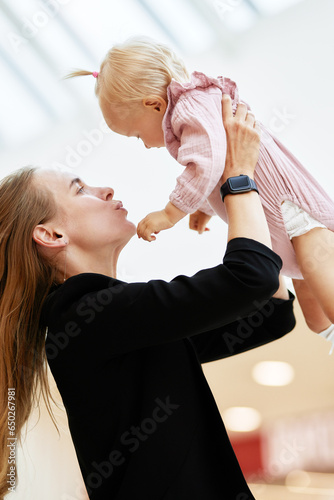 Young attractive mother lifts up her baby on outstretched arms in public place, shopping mall. Mom and little daughter in pink clothes play, relax and have fun while shopping. Family weekend in mall.