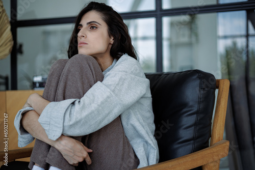 Disappointed brunette woman hugging knees and looking away while sitting on armchair at home