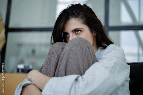 Frustrated brunette woman hugging knees and looking away while sitting at home