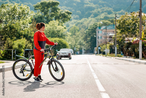 Sports and activity. Full-length portrait of a beautiful young woman in red sportswear, crossing the road on a Bicycle. Side view. In the background, a street and a road in the sunlight. Copy space