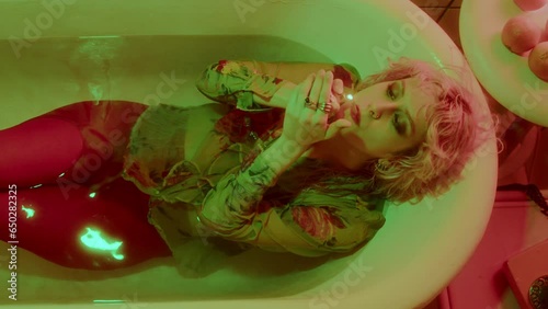 Young dressed girl with red lips lying in bathtub filled with water, lighting and smoking handrolled cigarette in bathroom with neon light. Top down shot photo