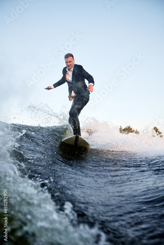 Young man surfer in classic suit rides on the wave. Happy clerk escaped from stuffy office to take up active sport. Best summer leisure after routine work. Film grain effect.