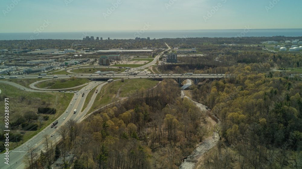A bird's eye view of a highway bridge over a valley with a creek and Lake Ontario in the background on a sunny spring day