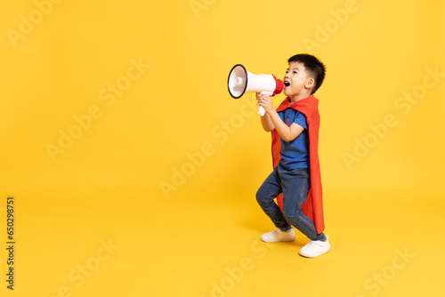 Superhero Asian boy with red cape holding megaphone isolated on yellow background, Wow and Announce Speech concept