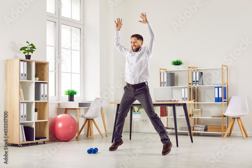 Happy funny fit smiling young business man or corporate employee doing star jumps during a quick fitness workout at work in the office. Sport, physical activity, healthy lifestyle concept